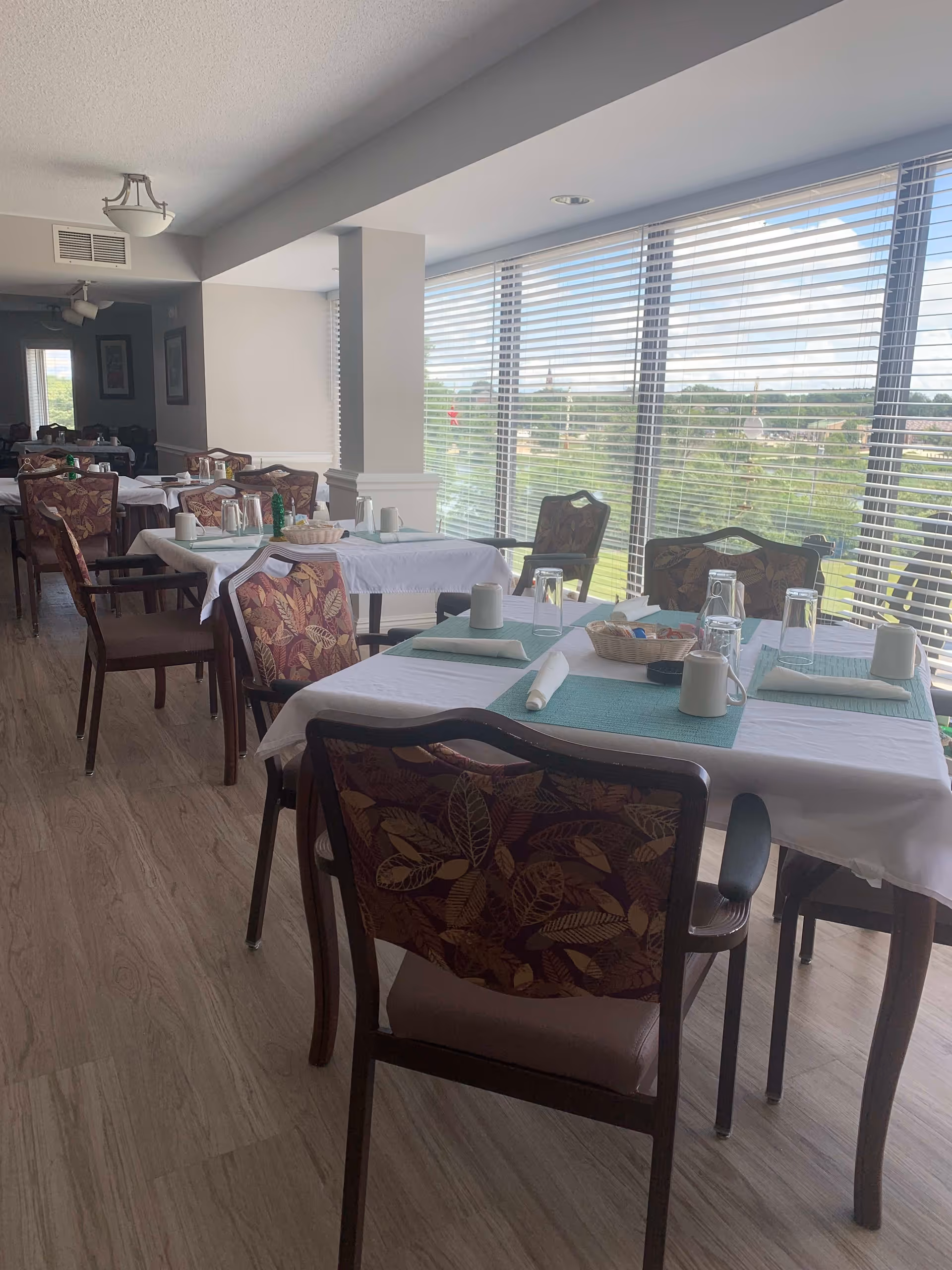 A dining room with several tables covered in white tablecloths and teal placemats. Each table is set with napkins, glasses, and cups. The chairs have a floral pattern on the backrests. Large windows with blinds allow natural light to fill the room and offer a view of greenery outside.