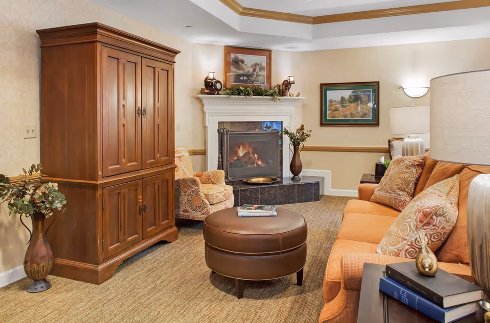 Cozy living room with a lit fireplace, orange sofa, patterned armchairs, a wooden armoire and a round leather ottoman.