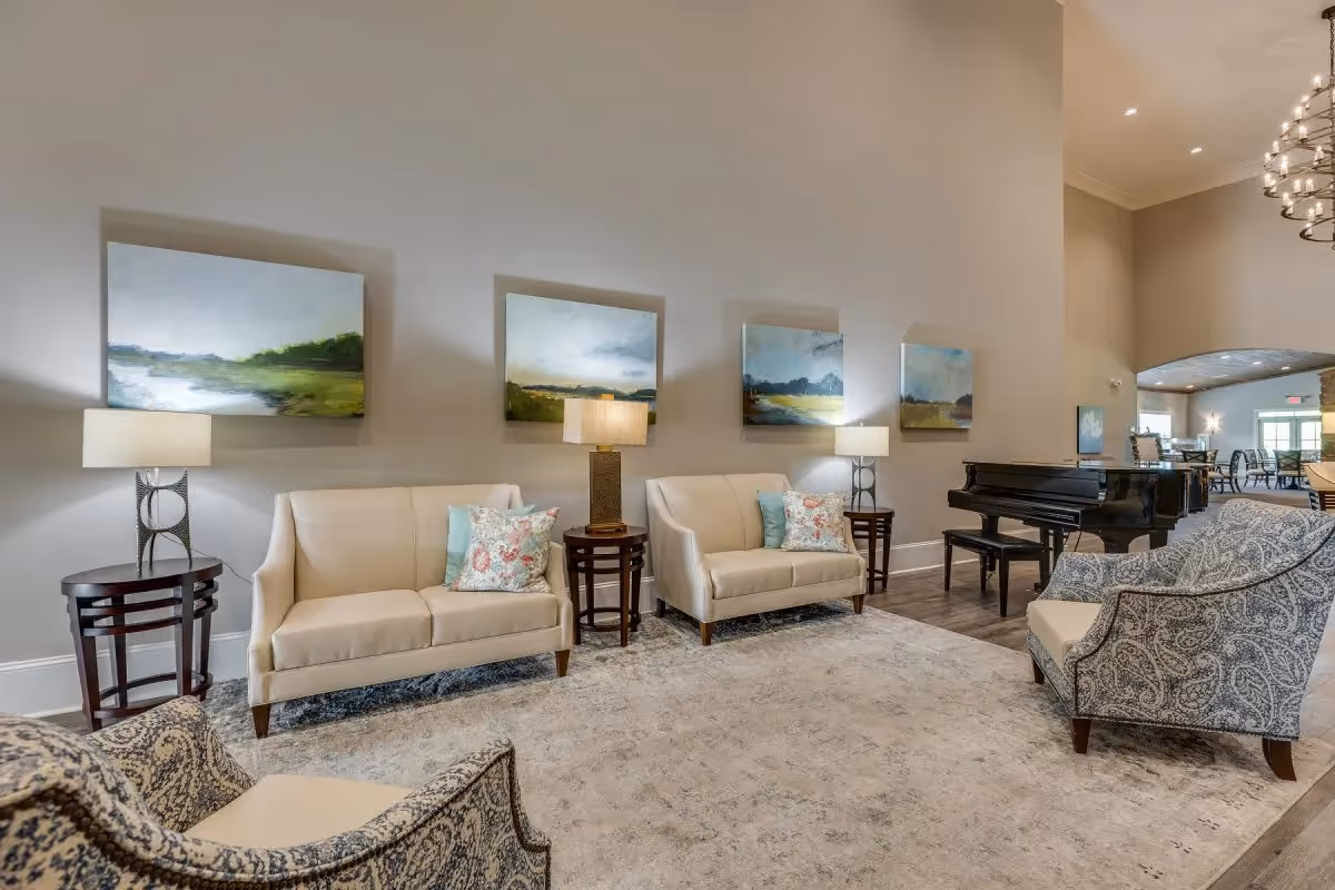 A spacious and well-lit living room area in an assisted living facility featuring two beige sofas with decorative pillows, two patterned armchairs, three side tables with lamps, a large area rug, and a black grand piano. The walls are adorned with four landscape paintings, and the room has high ceilings with recessed lighting and a chandelier.