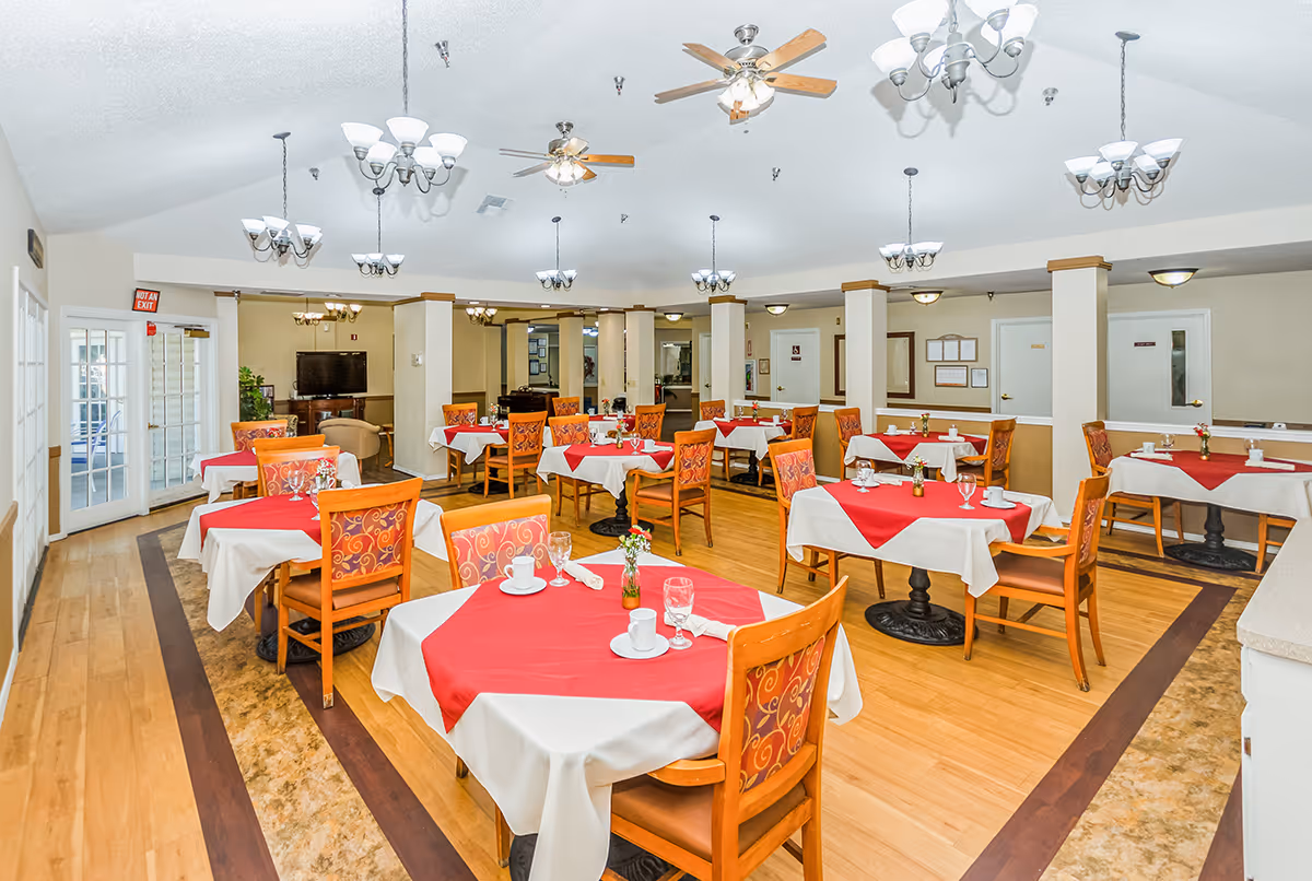 A dining room in a senior living facility with multiple tables covered in white tablecloths and red overlays. Each table is set with cups, glasses, napkins, and small flower vases. The room has wooden floors, ceiling fans, and multiple chandeliers providing lighting. There are several wooden chairs with patterned upholstery around the tables. In the background, there is a TV and some seating areas.