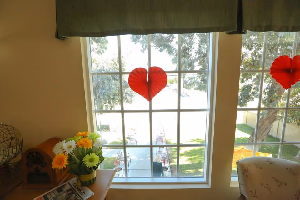 Interior view of a window decorated with red paper hearts, a small table with a vase of flowers and a chair, overlooking a sunlit lawn and patio.