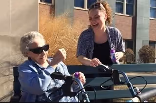 An elderly woman wearing sunglasses and a blue jacket is sitting outdoors with a walker in front of her, smiling and holding a small object. A younger woman standing nearby is also smiling and holding a purple flower. They are outside near a building with windows and some dry plants in the background.