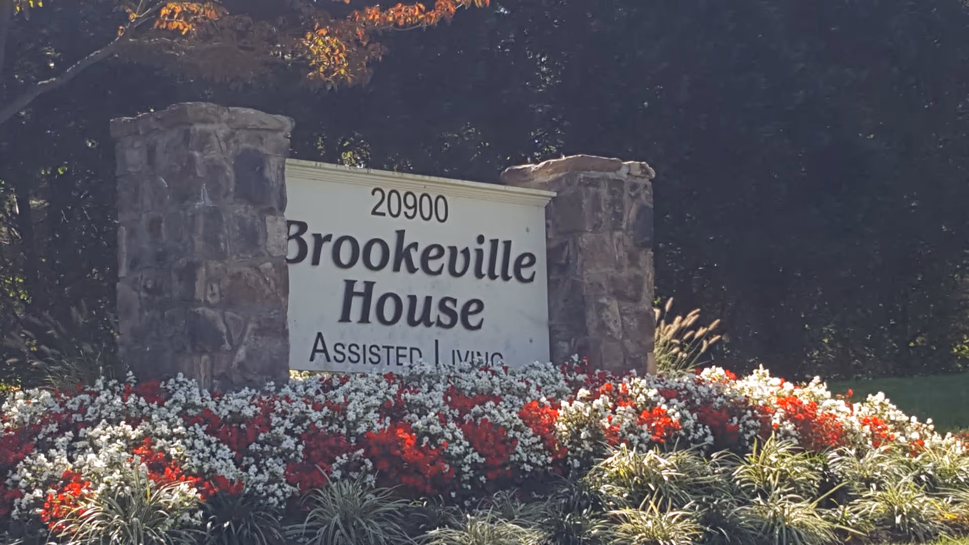 Stone signpost with the address 20900 and the name Brookeville House Assisted Living, surrounded by a flower bed with red and white flowers and greenery, with trees and bushes in the background.