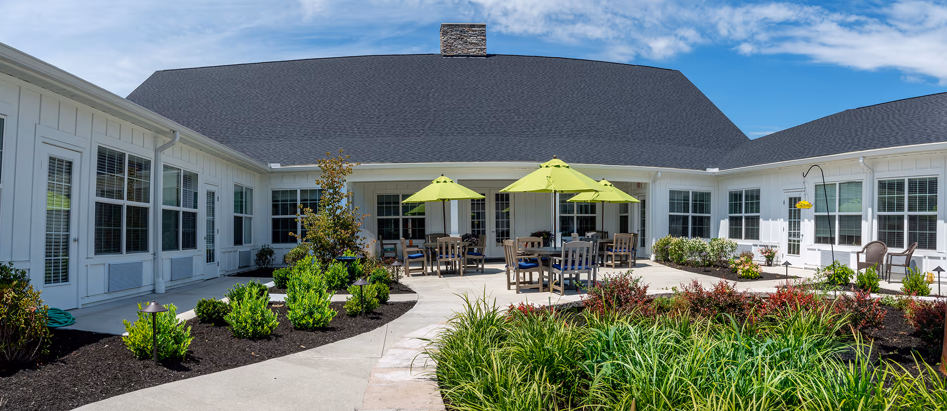 Outdoor patio area at Walnut Crossing featuring several tables with green umbrellas, surrounded by landscaped garden beds with various shrubs and plants, and white building walls with multiple windows and doors under a blue sky.