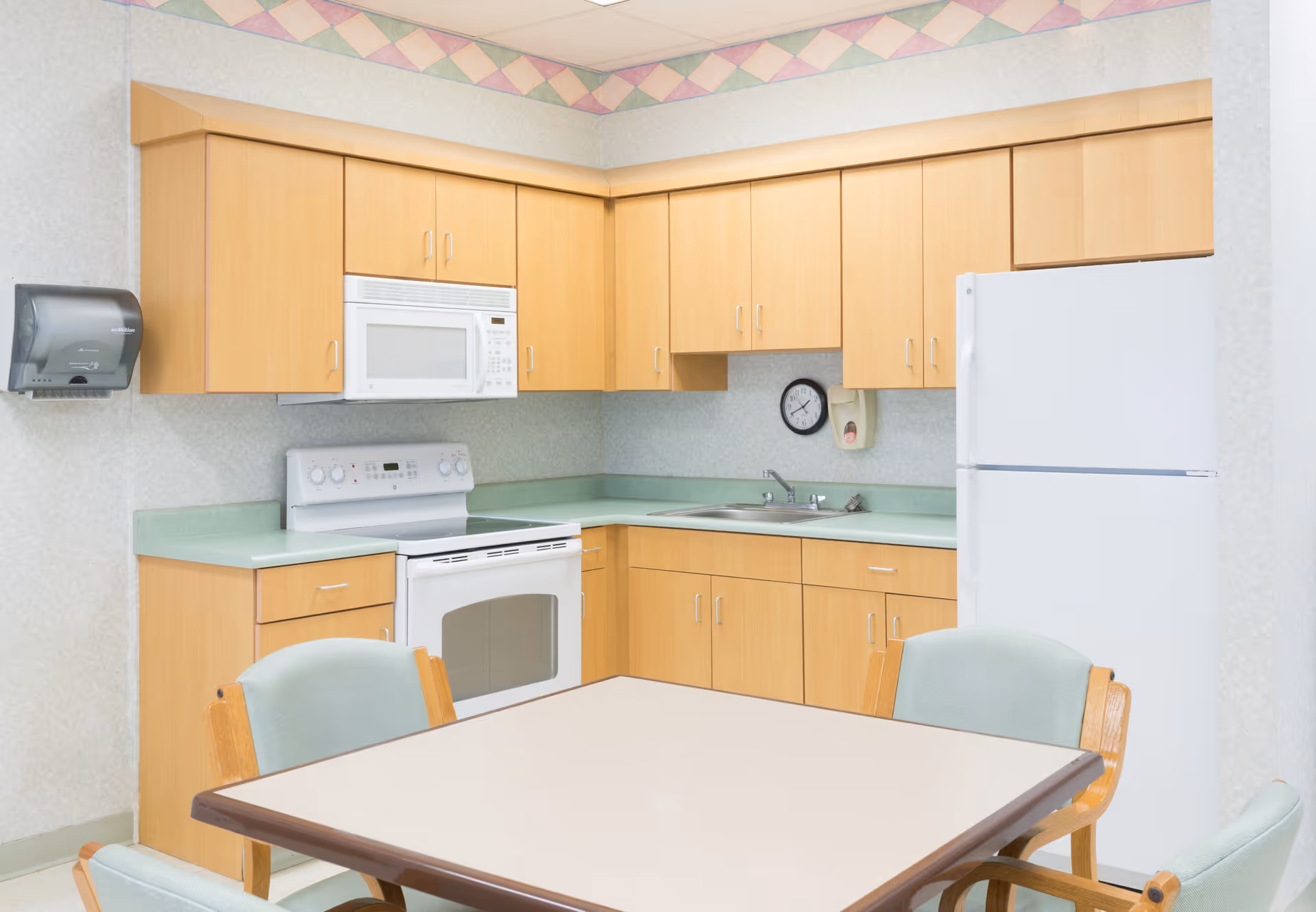 A small kitchen area with light wood cabinets, a white microwave above a white stove, a white refrigerator, a sink with a faucet, a wall clock, a soap dispenser, and a table with four chairs in the foreground. The walls have a light patterned wallpaper with a colorful geometric border near the ceiling.