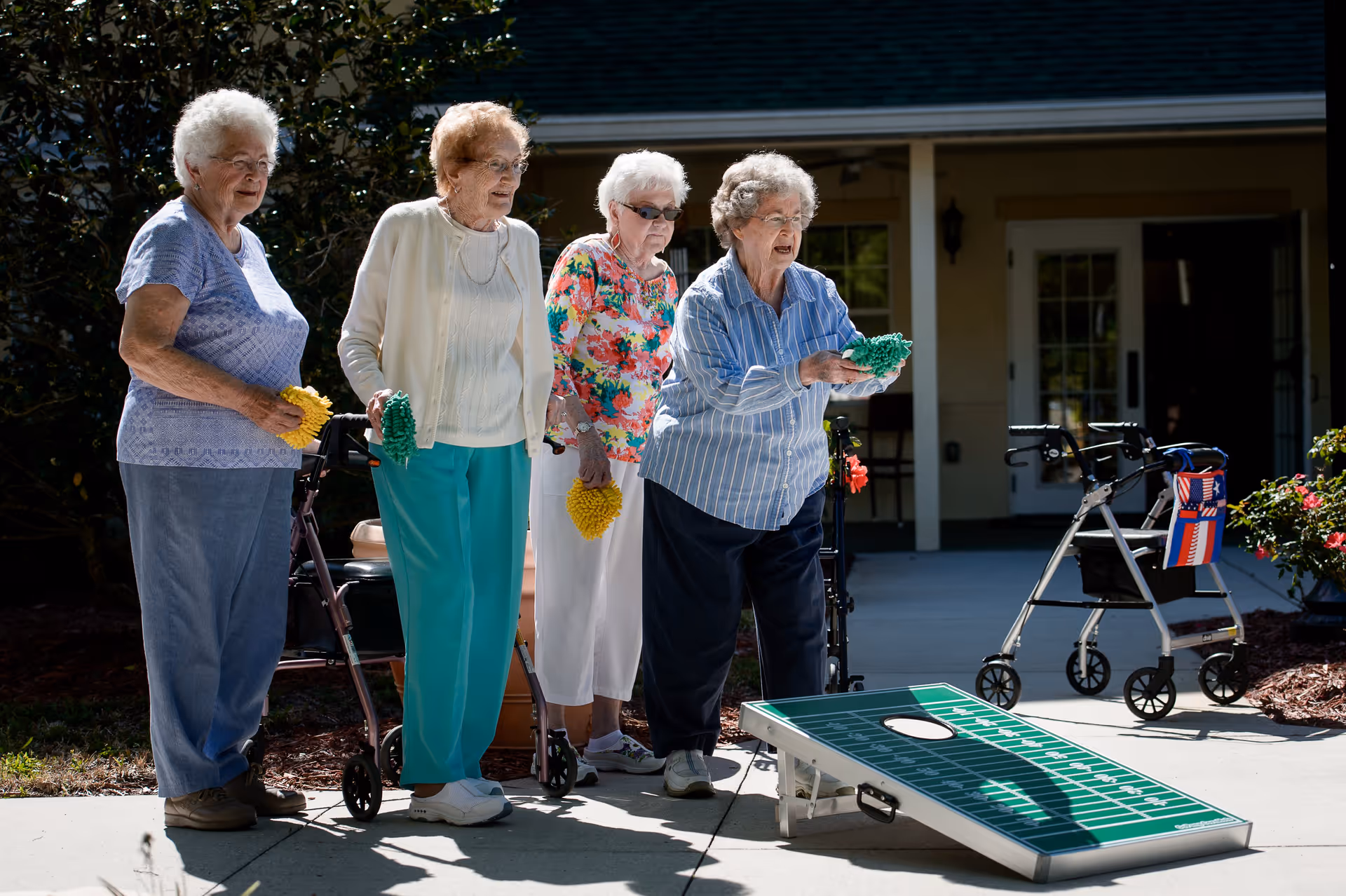 Four elderly women playing cornhole outdoors on a sunny day, each holding bean bags. Two walkers are visible nearby, and the setting appears to be a patio area outside a building.