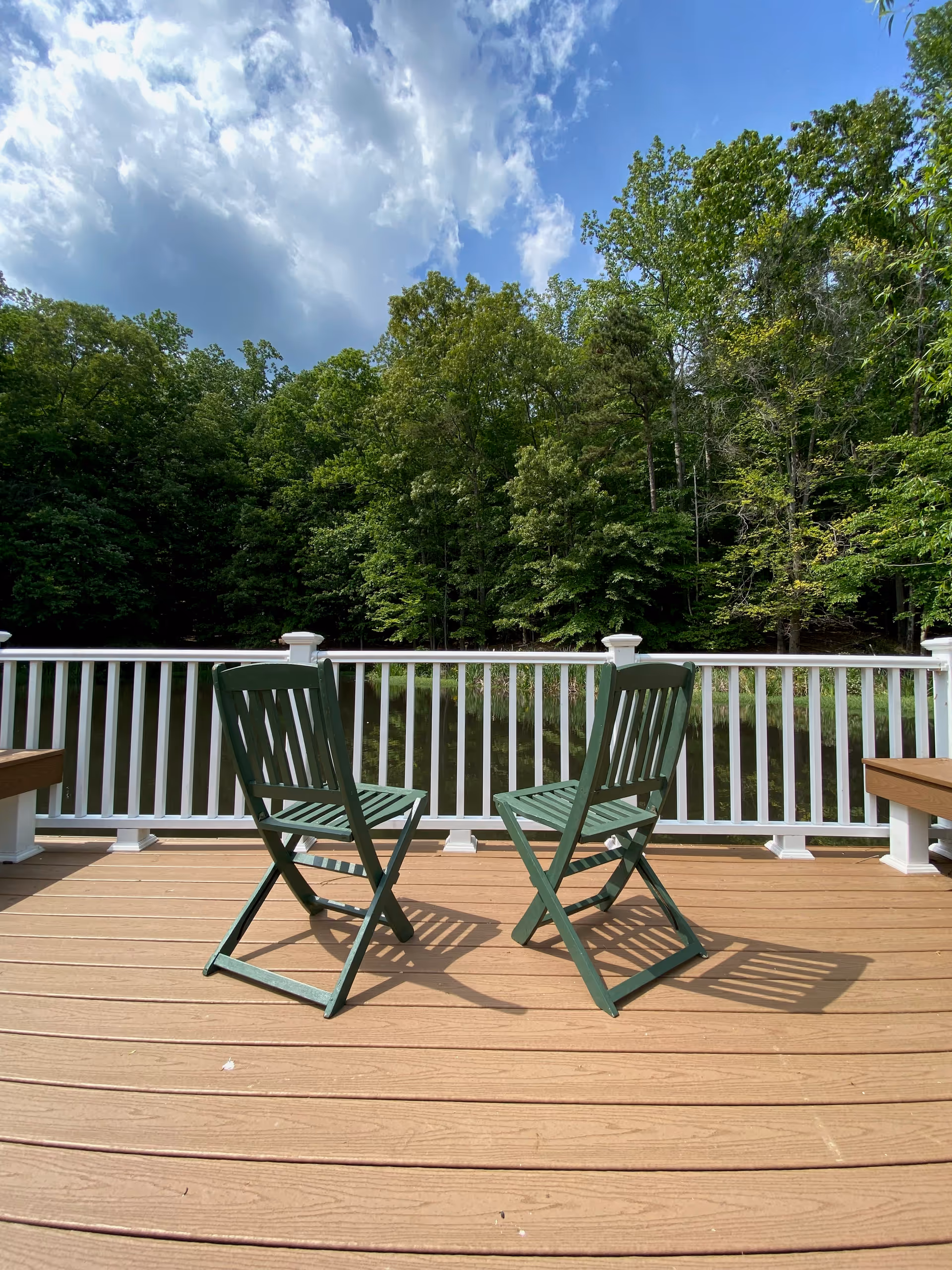 Two green wooden chairs on a wooden deck facing a white railing with a view of a pond and dense green trees under a partly cloudy blue sky.
