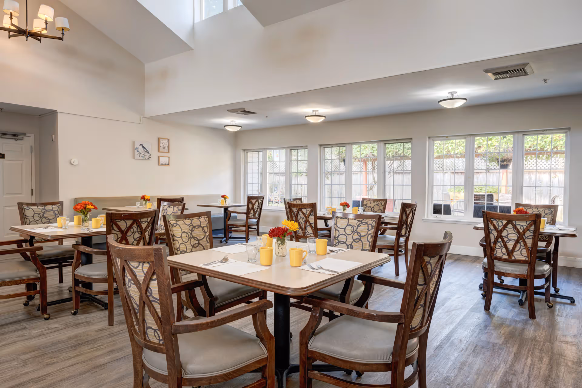 Bright dining room with multiple set tables and wooden chairs, large windows, and small floral centerpieces.