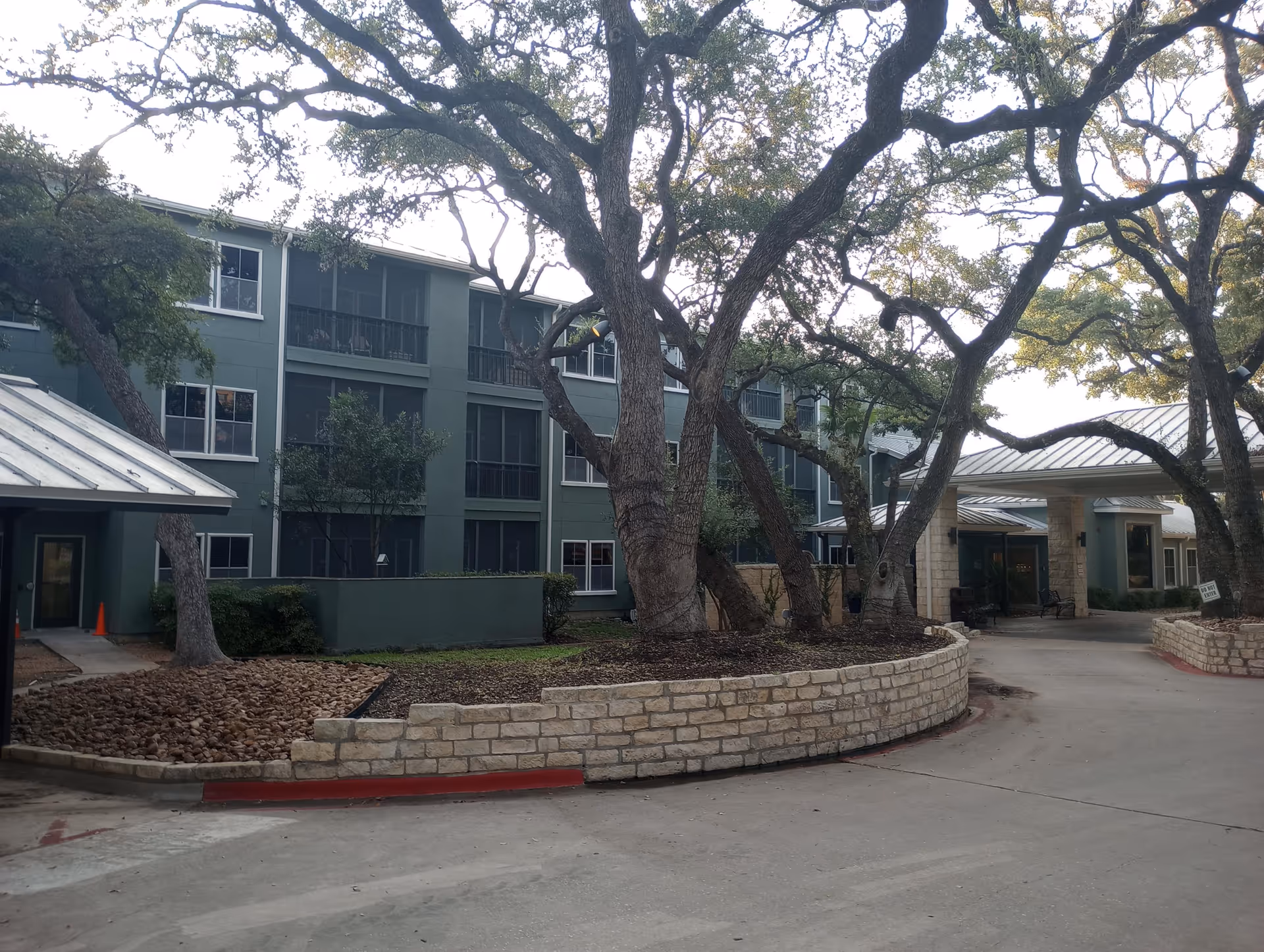 Front exterior of a multi-story senior living building with large oak trees, stone planters, and a covered entrance.
