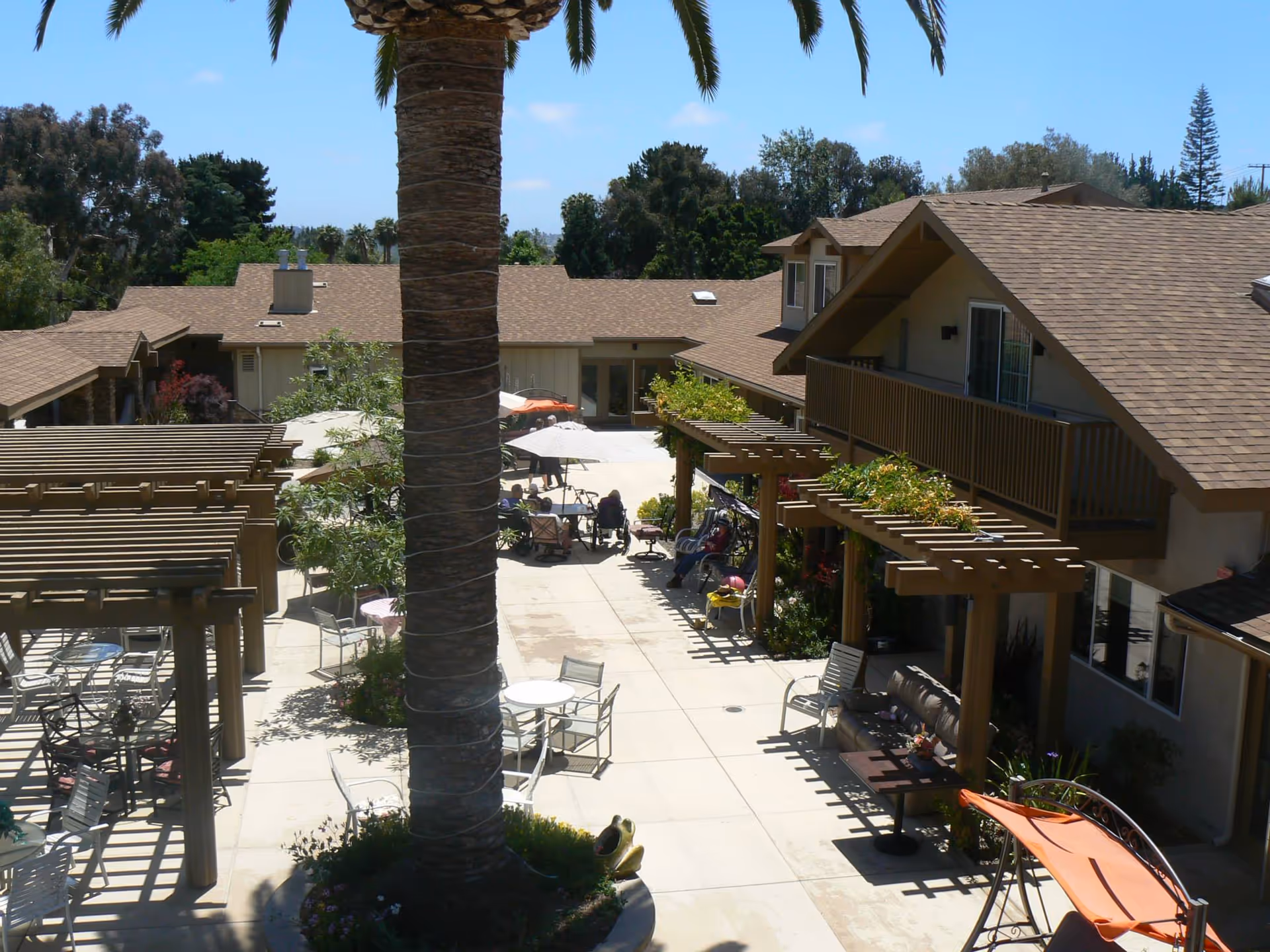 Sunlit courtyard with a central palm tree, pergolas, outdoor tables and chairs, and surrounding two-story buildings.
