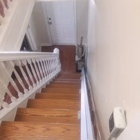 View looking down a wooden staircase with white railings inside a residential building, showing a closed white door at the bottom and a stair lift attached to the right side of the stairs.