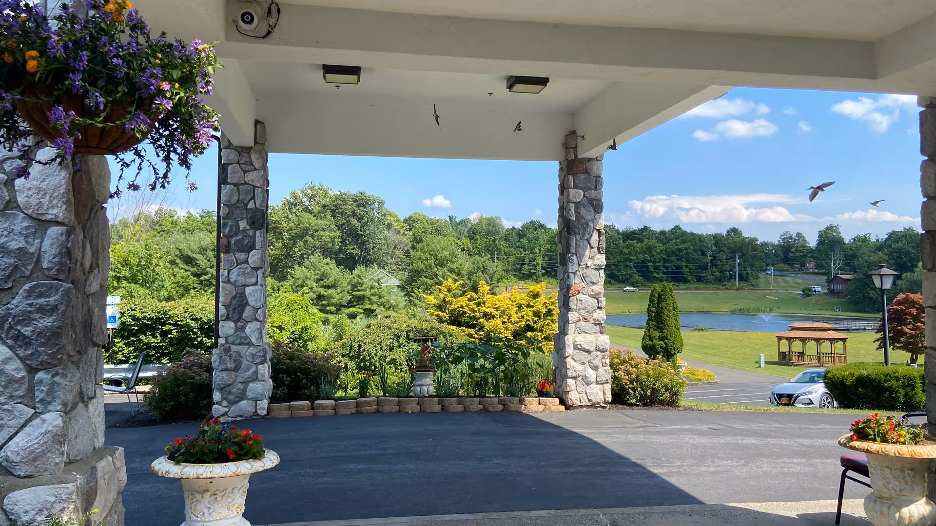 View from a covered stone entrance area looking out onto a landscaped garden with green trees, shrubs, and a small pond with a gazebo. There are hanging flower baskets and potted plants near the entrance, with a few cars parked in the background under a blue sky with some clouds.