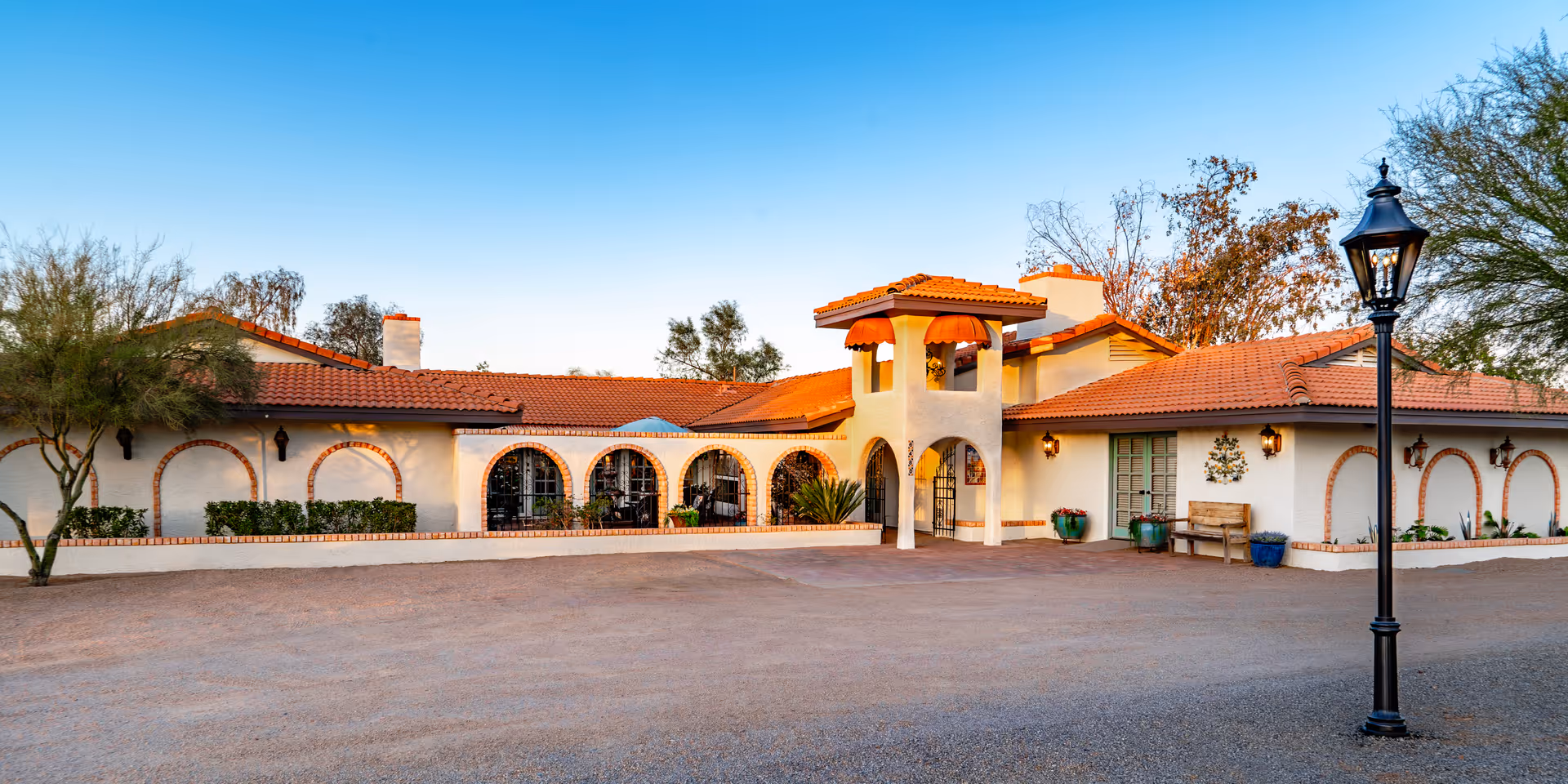 Exterior view of a single-story assisted living facility with white walls and a red tiled roof. The building features arched windows and doorways, decorative brick accents, and a small tower-like structure above the entrance. There are potted plants and a wooden bench near the entrance, with a black street lamp in the foreground and trees surrounding the building under a clear blue sky.