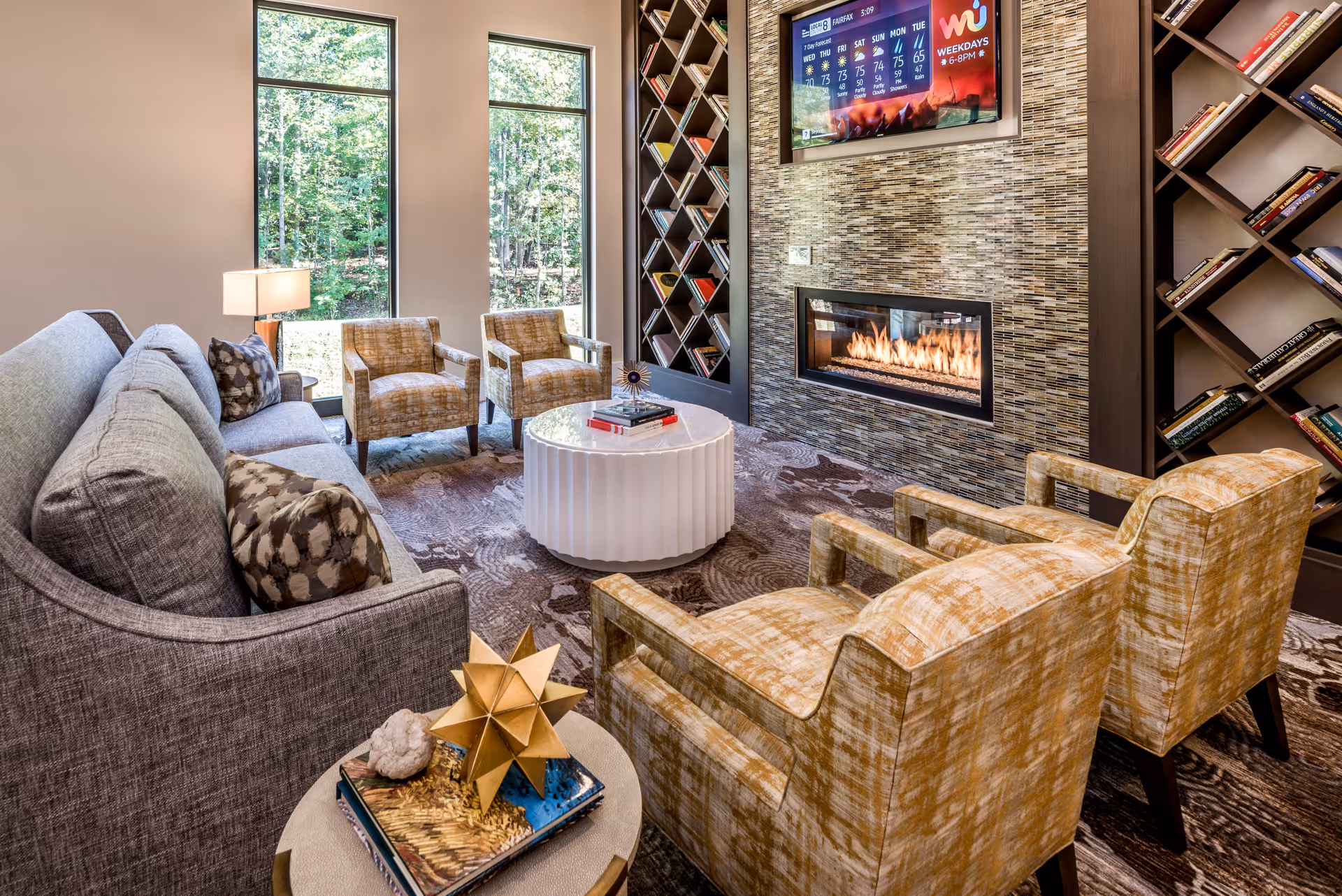 A cozy living room area with a gray sofa, four patterned armchairs, a round white coffee table, and a modern fireplace set in a tiled wall. Two tall windows let in natural light and show a view of green trees outside. Bookshelves with books flank the fireplace, and a flat-screen TV above the fireplace displays a weather forecast.