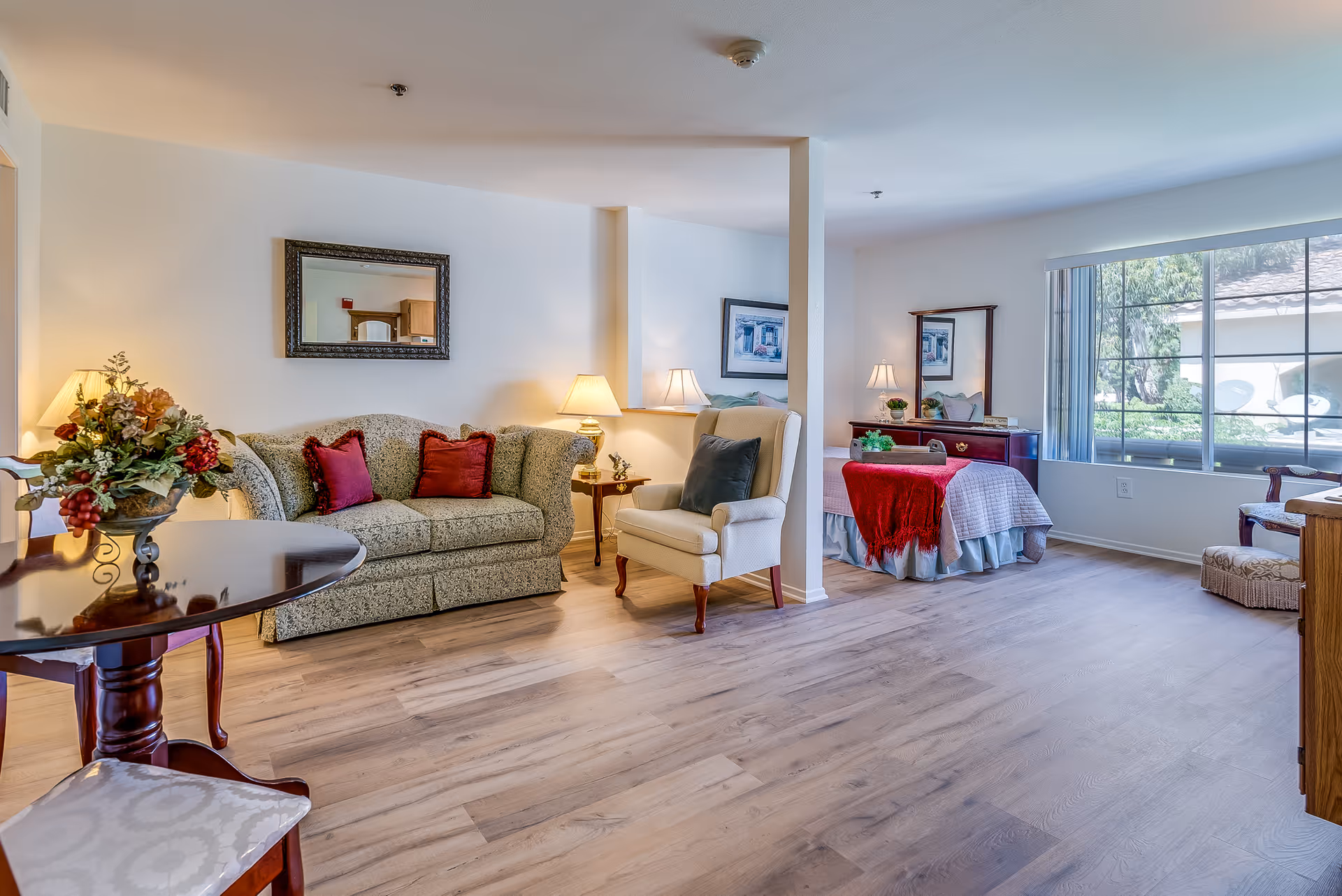 Bright open living area with a sofa, armchair, side tables and a bed by a large window in a senior assisted living suite.