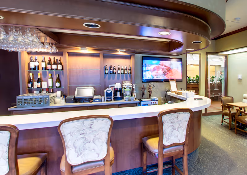 Interior view of a bar area with a curved white countertop and wooden bar stools with floral upholstery. Behind the bar, there are shelves stocked with various bottles of wine and liquor, a coffee machine, and other beverage equipment. A flat-screen TV is mounted on the wall displaying a program. To the right, there is a glass door leading to another room with chairs and tables.