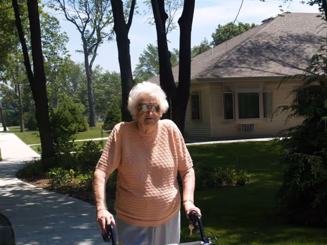 An elderly woman with white hair wearing sunglasses and a peach-colored shirt is walking outdoors on a paved pathway using a walker. There are trees, grass, and a building with windows in the background under a clear sky.