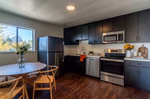 Modern kitchen with dark cabinets, stainless steel refrigerator, dishwasher, oven, and microwave. A round wooden dining table with four chairs is placed near a window with a vase of flowers on the table. The floor is wooden and the kitchen is well-lit with a ceiling light.