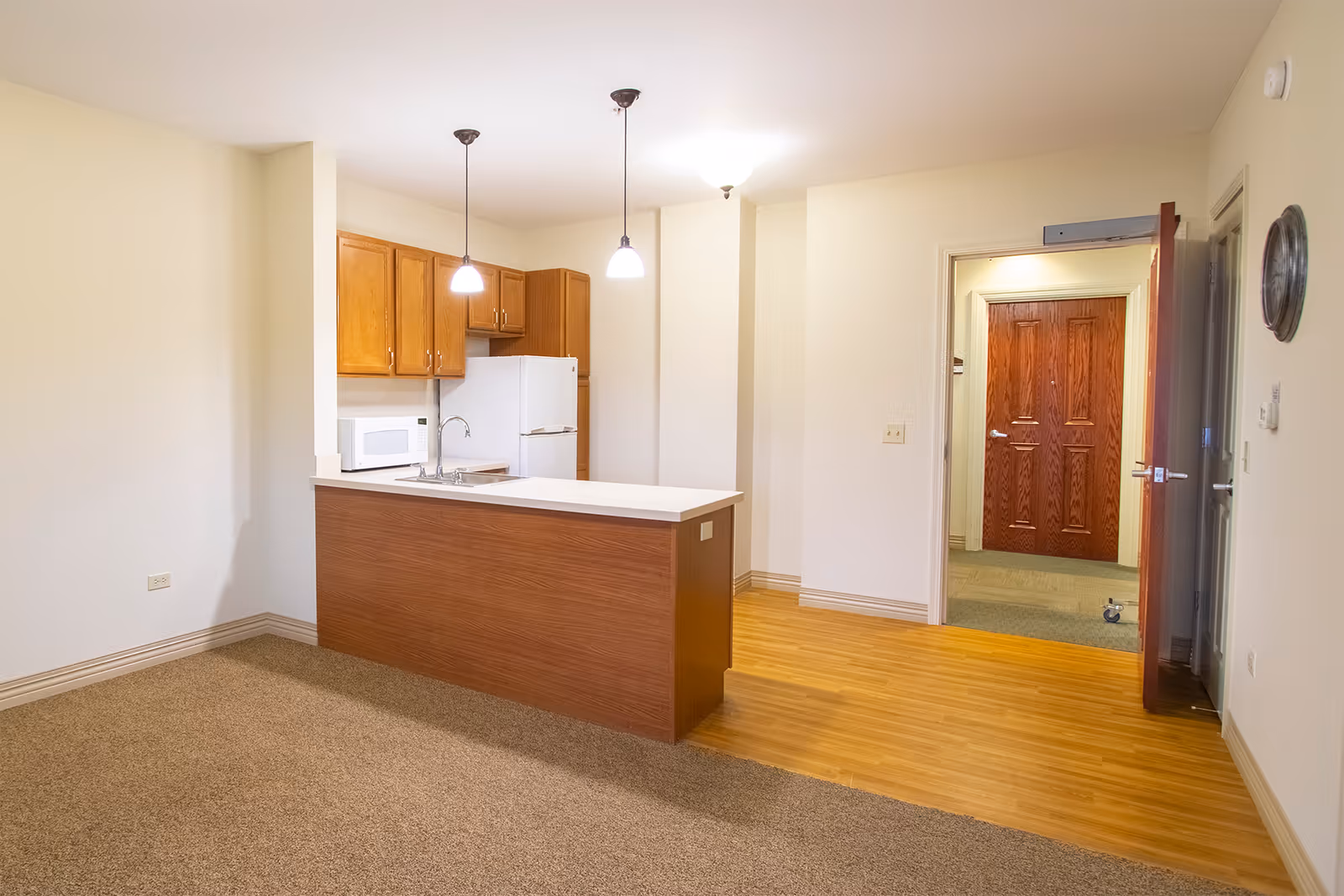 Interior view of a senior living facility apartment kitchen area with wooden cabinets, a white refrigerator, a microwave, and a countertop with a sink. The floor transitions from carpet in the living area to wood laminate in the kitchen. There are two pendant lights hanging over the counter and a ceiling light near the entrance door, which is open showing a hallway with a wooden door.