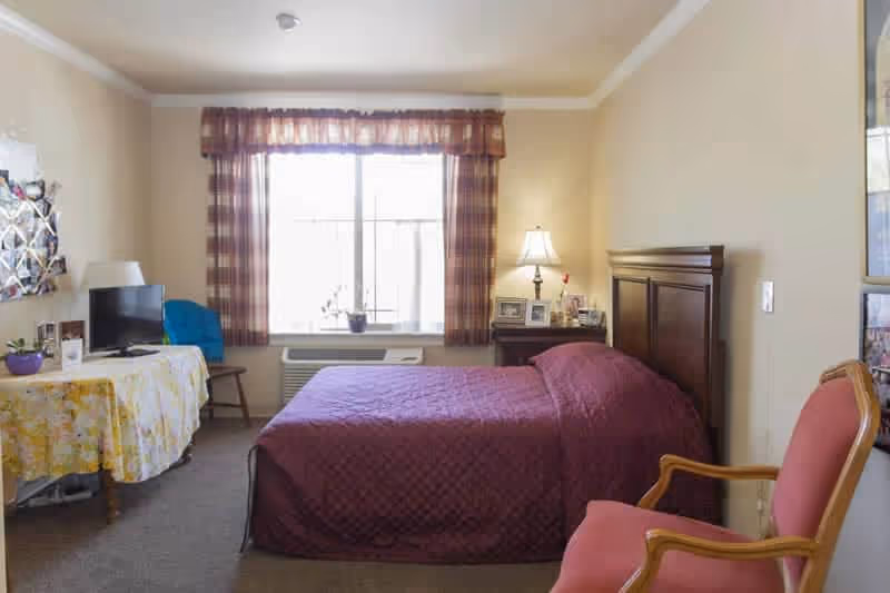 A cozy bedroom with a double bed covered in a maroon quilt, a wooden headboard, and a bedside table with a lamp and framed photos. There is a window with plaid curtains letting in natural light, a table covered with a floral tablecloth holding a small TV and other items, and a pink upholstered chair with wooden arms.