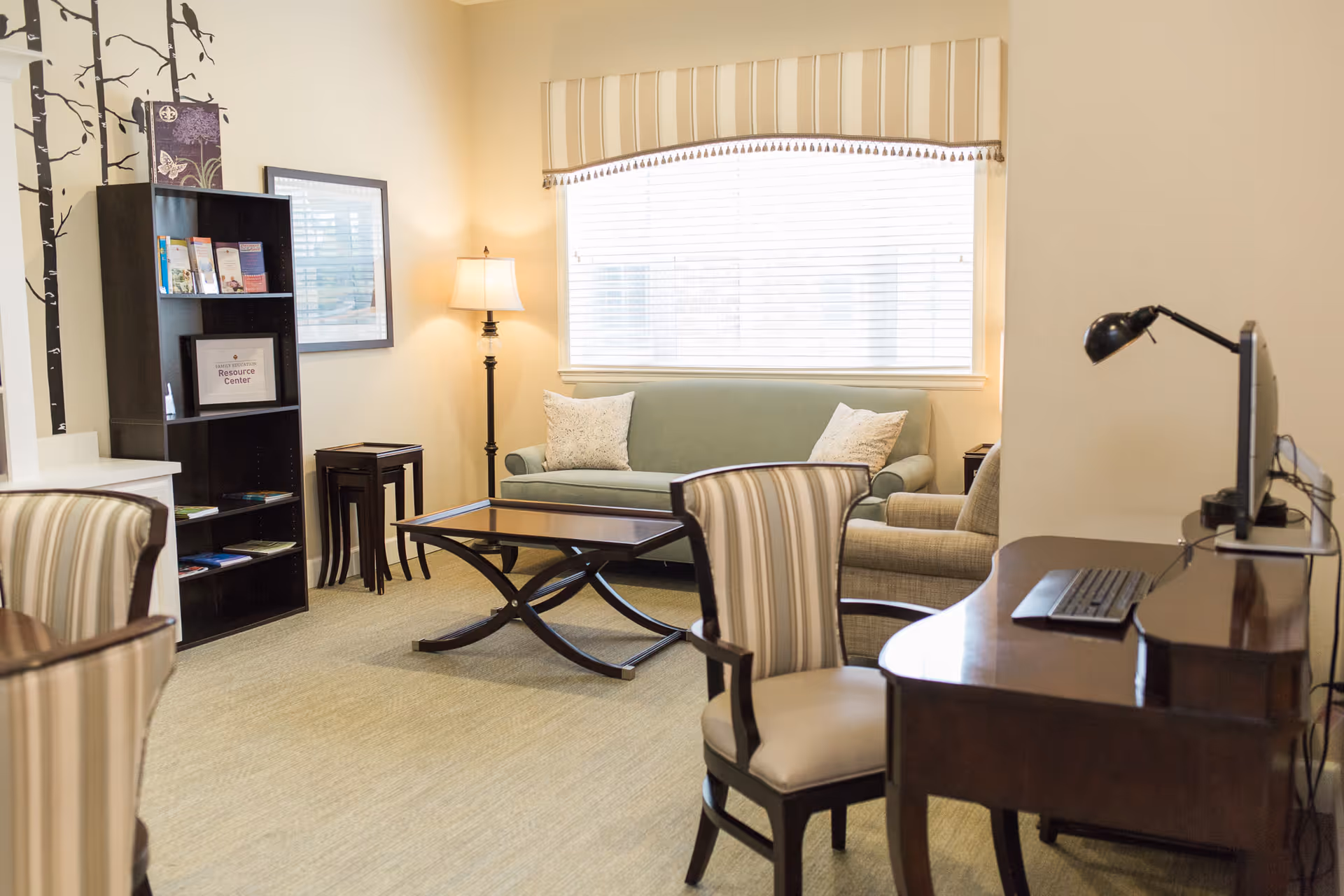 A well-lit living room area with a green sofa, two armchairs, a wooden coffee table, a floor lamp, a bookshelf with brochures, and a desk with a computer and lamp. The room has a large window with blinds and a valance, and the walls are painted a light beige color.