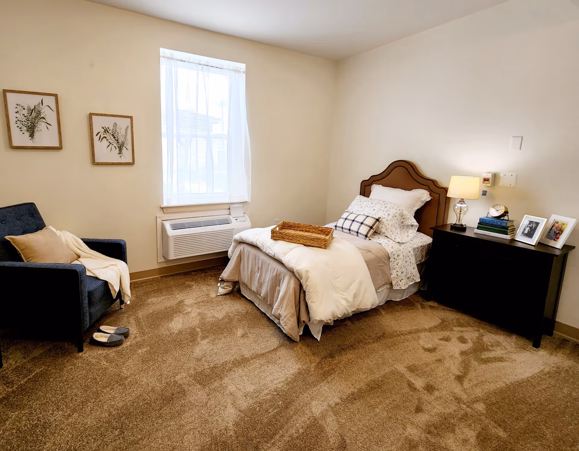 A cozy bedroom with a single bed featuring a brown headboard, white and patterned bedding, and a wicker tray on top. Next to the bed is a black nightstand with a lamp, books, a clock, and framed photos. A blue armchair with a beige pillow and cream throw blanket is positioned near a window with sheer white curtains. The room has beige carpet and light-colored walls with two framed botanical prints hanging on one wall.