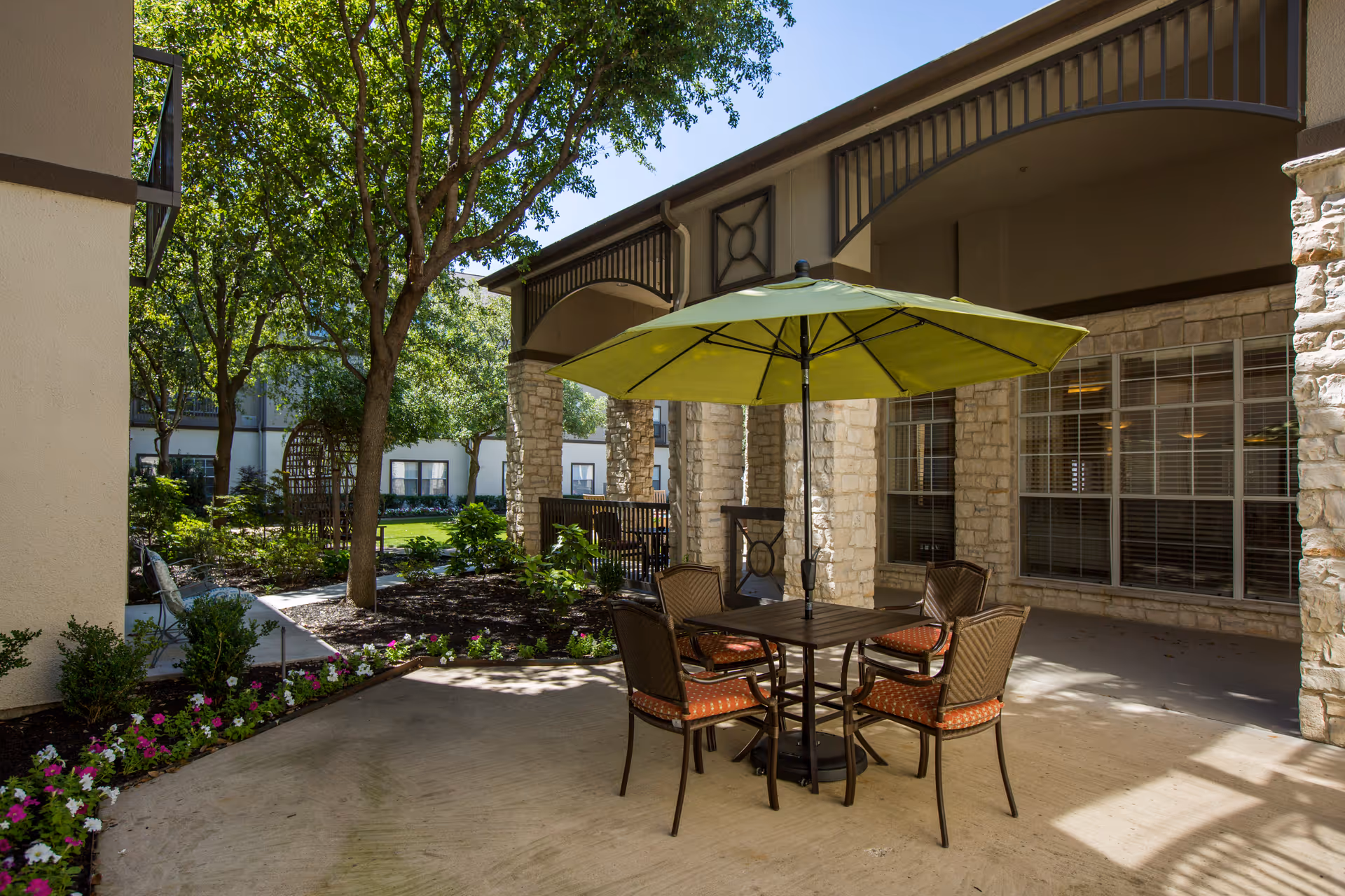Outdoor patio area with a square table and four chairs under a large green umbrella. The patio is surrounded by stone columns and overlooks a garden with trees and flowers. The building exterior features large windows with blinds.