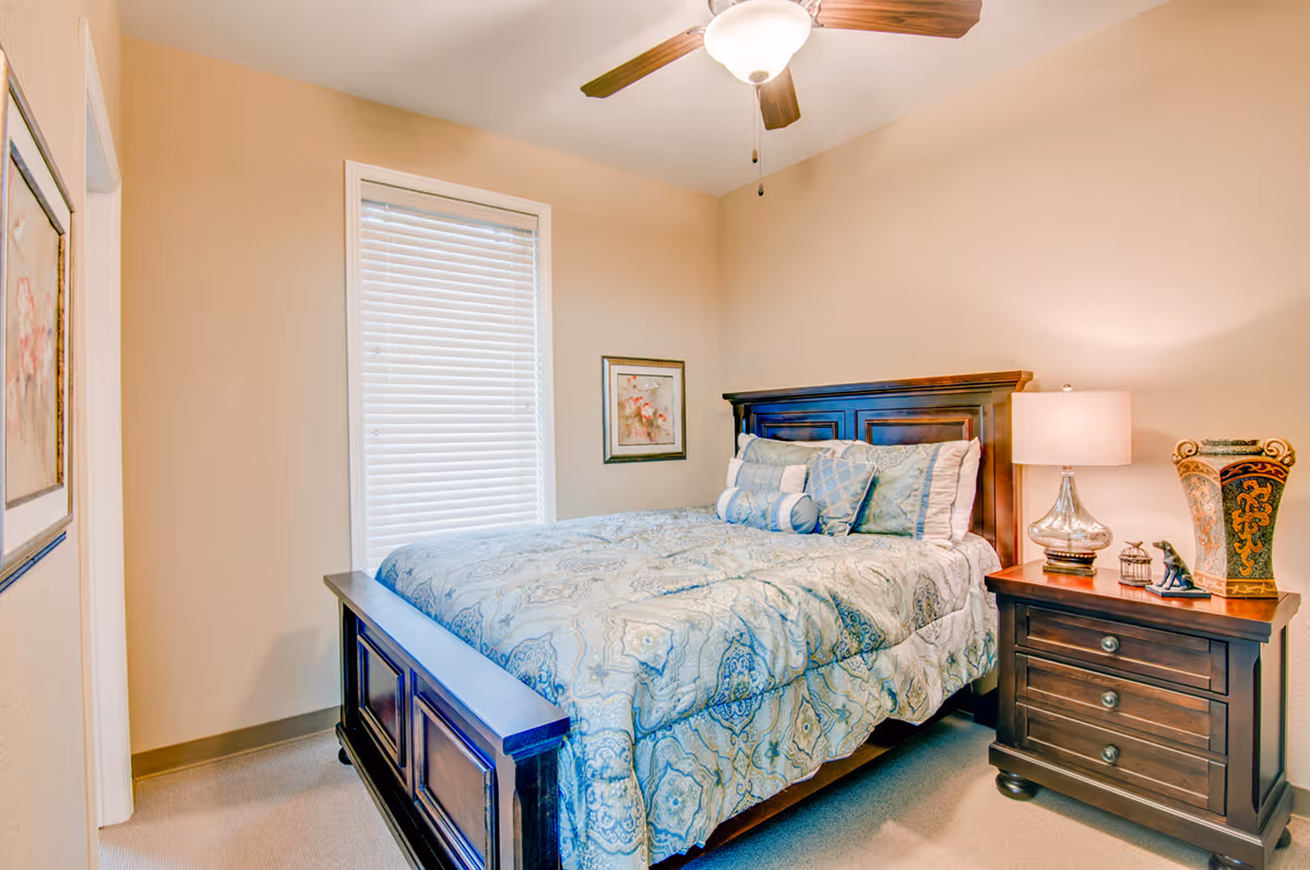 A cozy bedroom with a wooden bed frame and a bed made with a blue patterned comforter and multiple pillows. There is a wooden nightstand with a lamp, decorative vase, and small figurines. The room has beige walls, a window with white blinds, and framed artwork on the walls.