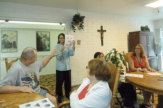 A group of elderly people seated around a wooden table in a room with white brick walls. A woman standing is holding up a paper with shapes on it, seemingly leading an activity or discussion. The room has plants, a crucifix on the wall, and framed pictures in the background.