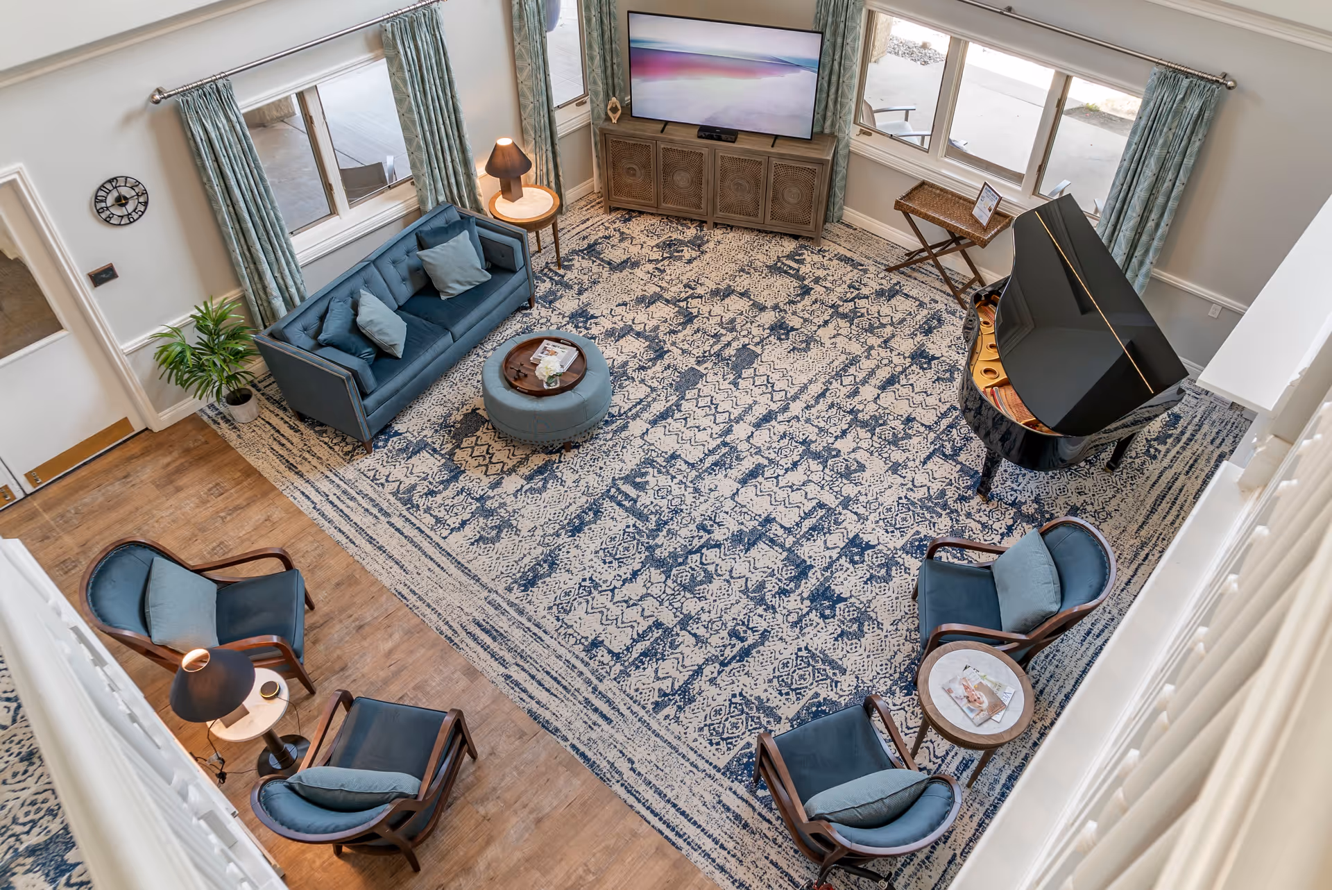 A spacious living room with a large patterned area rug, a blue sofa with cushions, four blue cushioned chairs arranged around small round tables, a black grand piano, a wooden cabinet with a flat-screen TV on top, large windows with light blue curtains, and a potted plant near the door.
