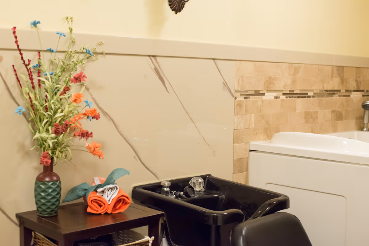A salon area with a black hair washing sink and chair next to a wooden table holding a vase with colorful artificial flowers and neatly folded towels. The background features beige marble and tile walls.