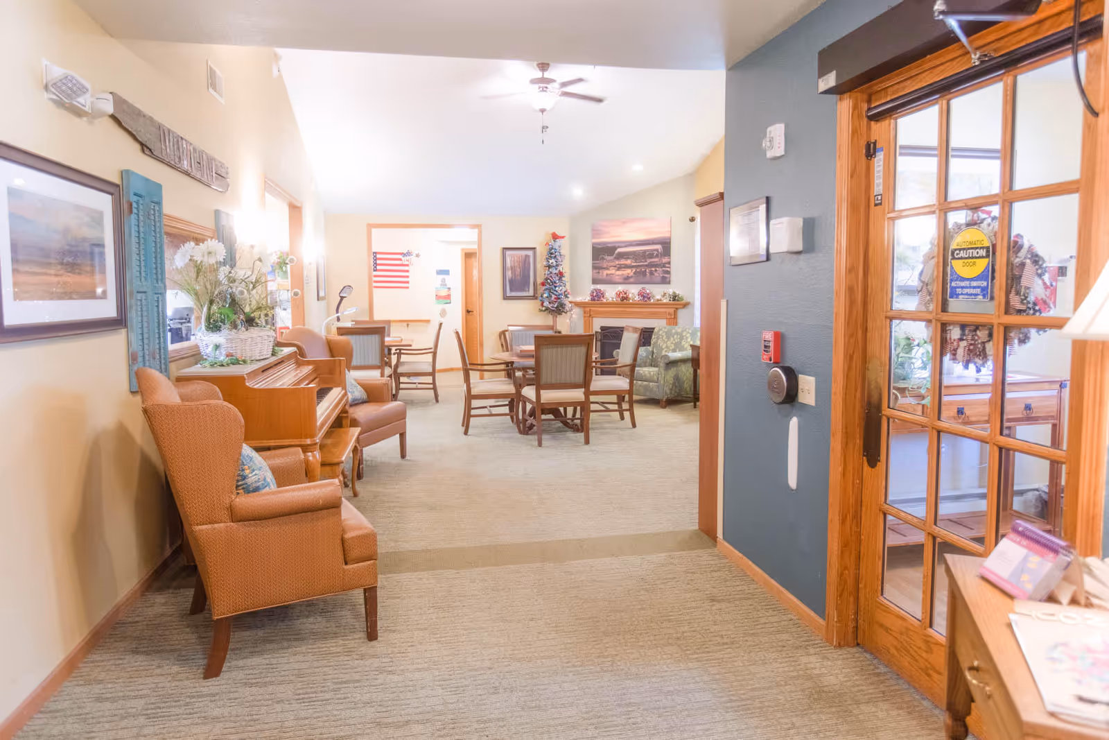 Well-lit assisted living common lounge with armchairs, a piano, and tables and chairs visible through a glass-paneled entrance door.