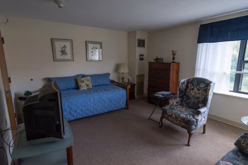 A cozy living room with a blue quilted daybed against the wall, two framed botanical prints above it, a wooden side table with a lamp, a wooden chest of drawers, a floral upholstered armchair, a small folding table with a book, and a window with sheer white curtains and a blue valance letting in natural light.