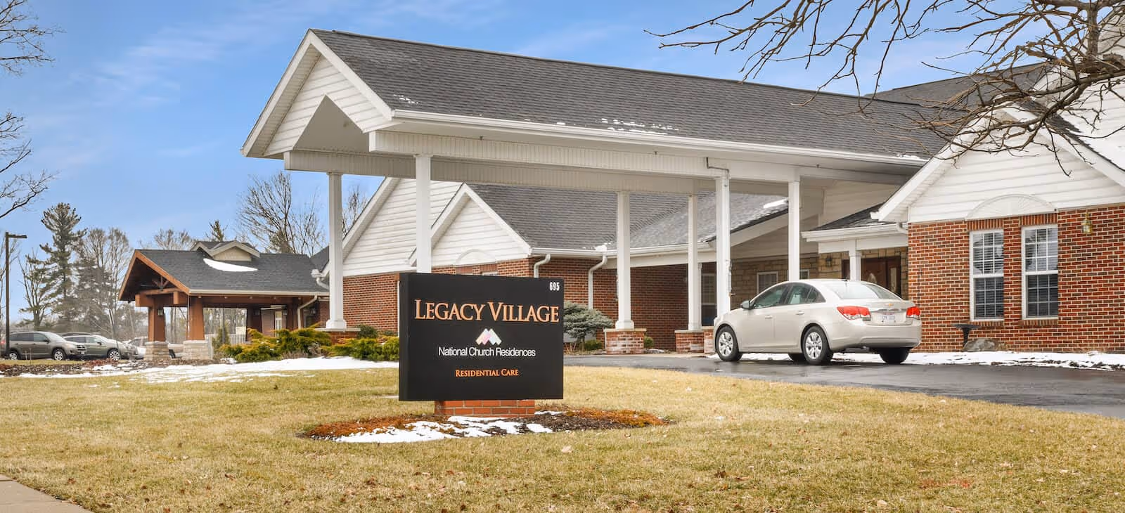 Exterior view of Legacy Village, a residential care facility with a brick building and white trim. A covered driveway with a white car parked under it is visible. There is a sign in front that reads 'Legacy Village National Church Residences Residential Care'. The lawn has patches of snow and leafless trees are visible.