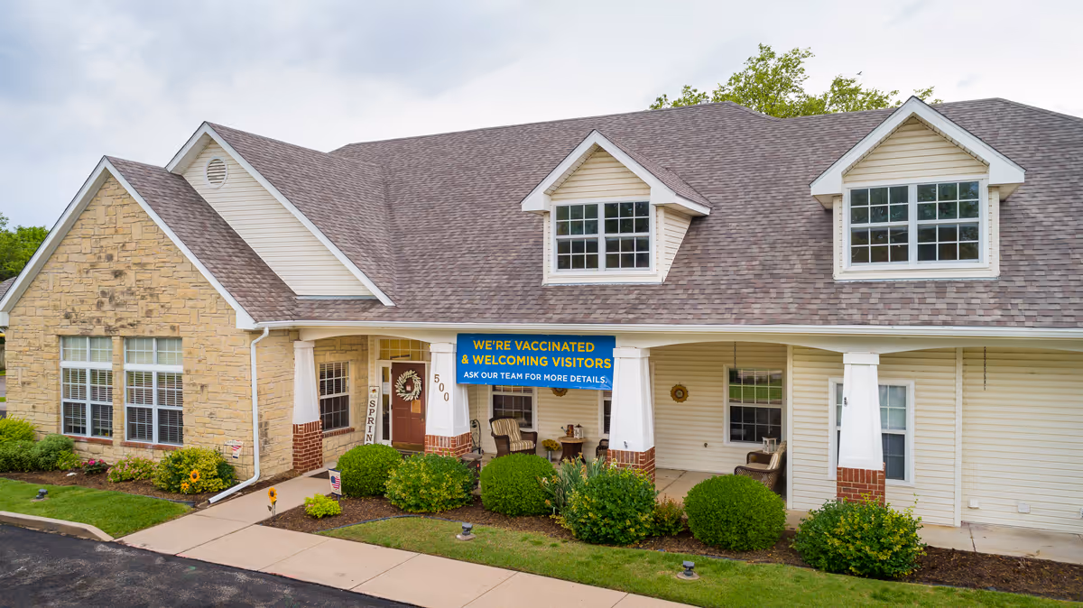 Front exterior of an assisted living building with a covered porch, landscaping, and a banner reading "We're vaccinated & welcoming visitors."