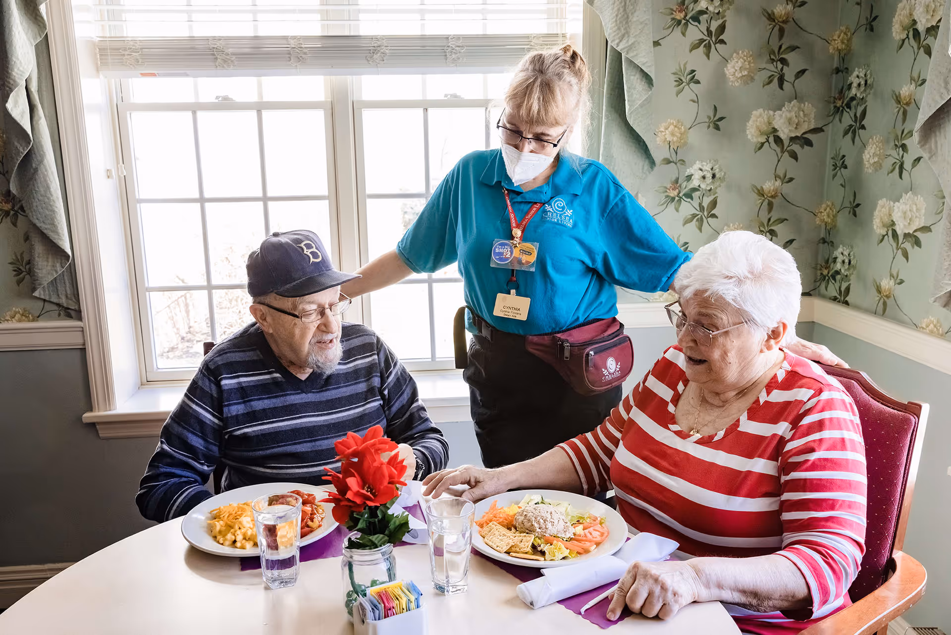 An elderly man and woman sit at a dining table with plates of food and glasses of water. A caregiver wearing a mask and a blue shirt stands behind them, engaging warmly with the couple. The room has floral wallpaper and a large window letting in natural light.