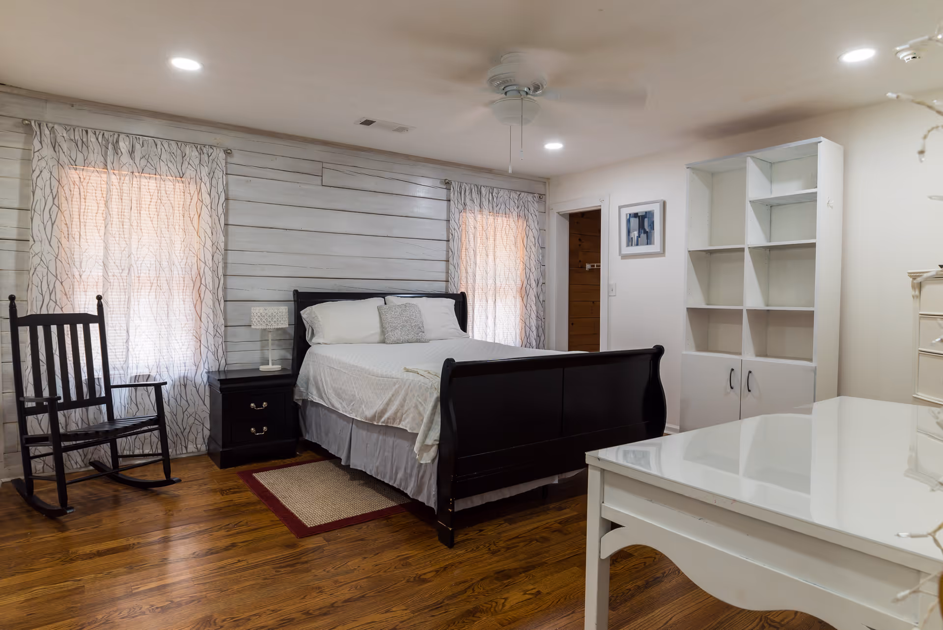 Well-lit bedroom with a dark wood sleigh bed, rocking chair, white shelving and desk, and hardwood floors.