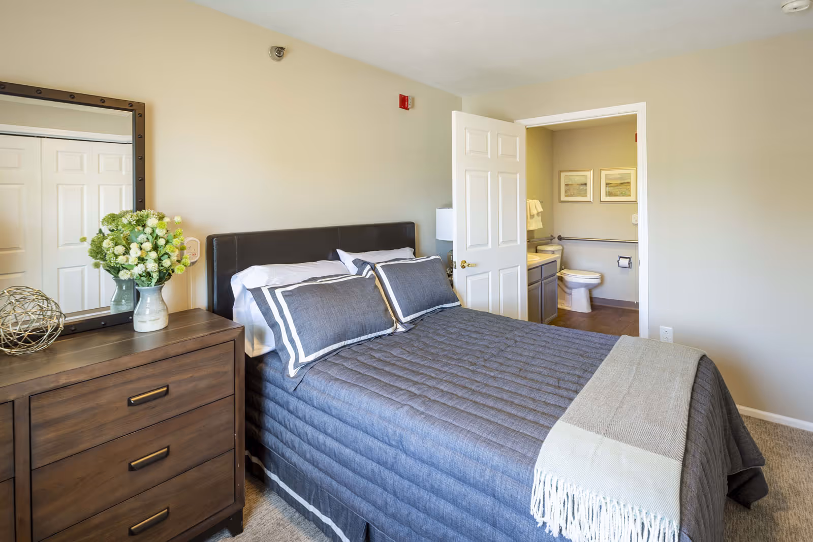 A bedroom with a neatly made bed featuring gray bedding and pillows. To the left of the bed is a wooden dresser with a large mirror above it, decorated with a vase of flowers and a decorative object. An open door reveals a bathroom with a toilet, sink, and two framed pictures on the wall.