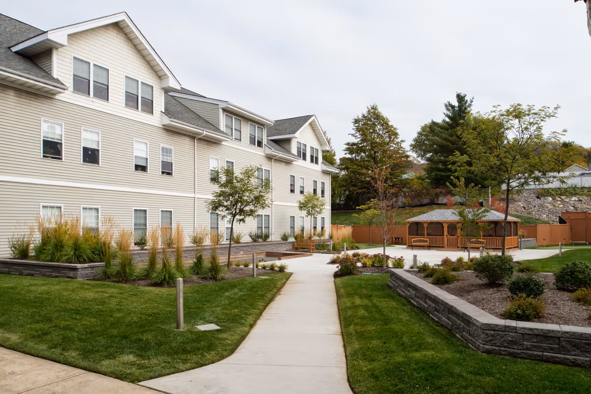 Exterior view of a multi-story assisted living facility with beige siding, multiple windows, and a paved walkway leading to a wooden gazebo surrounded by landscaped greenery and trees.