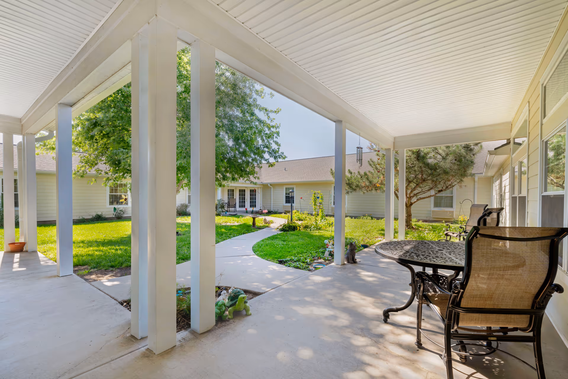 Covered outdoor patio area with a round metal table and chairs, overlooking a green courtyard with a concrete pathway, trees, and a beige building in the background.