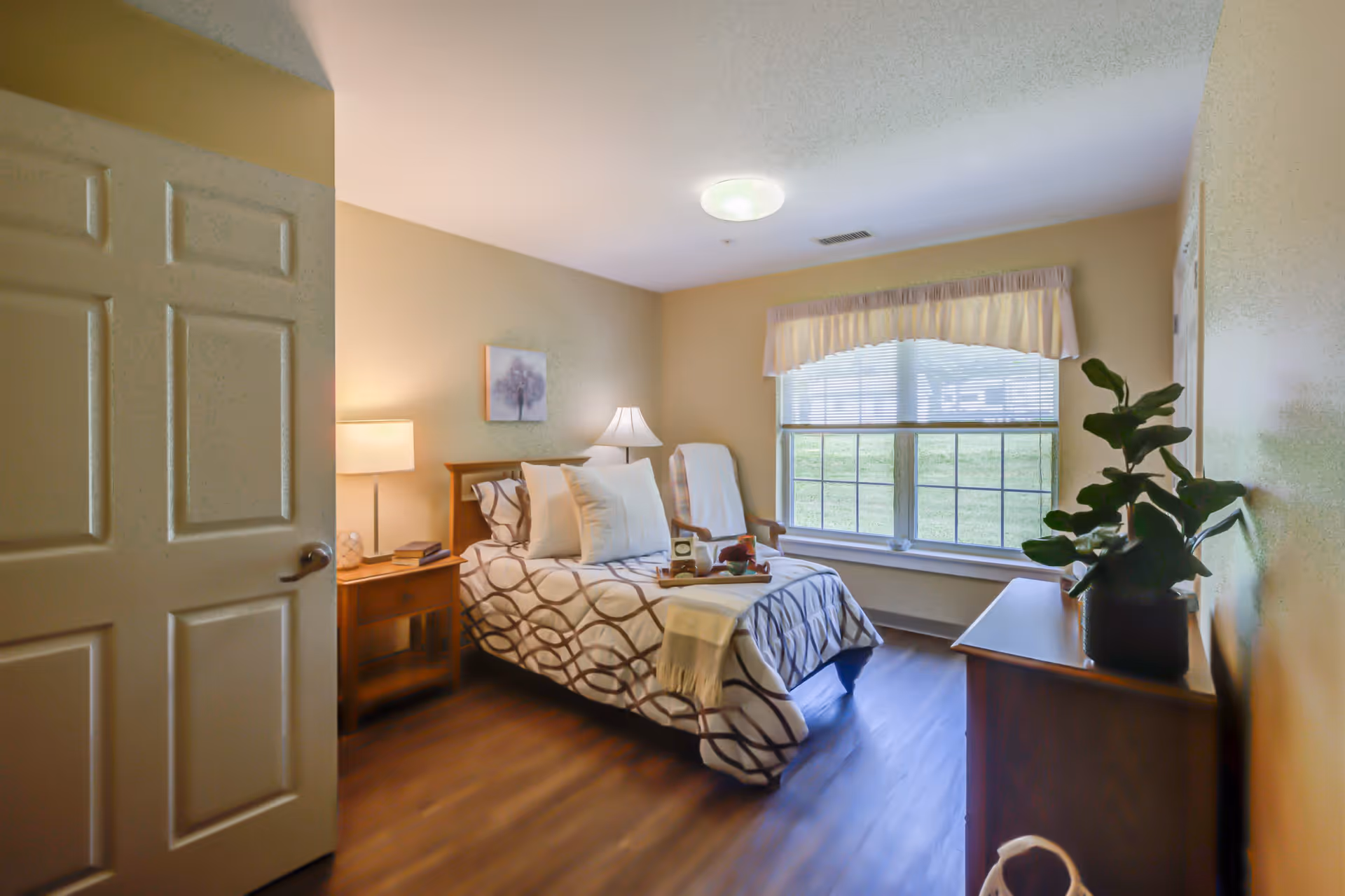 A cozy bedroom in a senior living facility featuring a single bed with patterned bedding and multiple pillows. There is a wooden nightstand with a lamp and books beside the bed. A comfortable armchair with a white cover is placed near a large window with blinds and a valance. A wooden dresser with a potted plant sits against the opposite wall. The room has warm lighting and wood flooring.