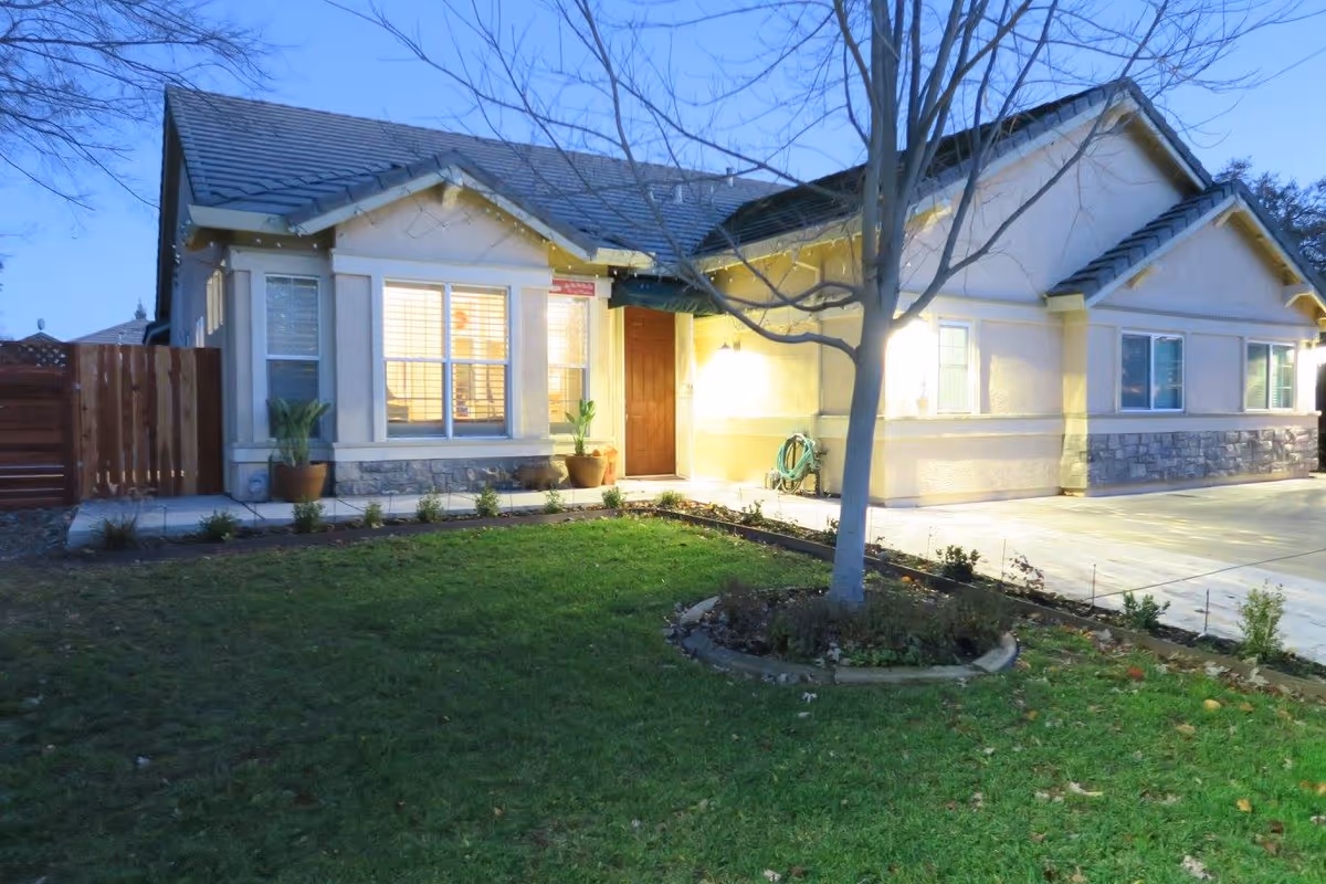 Front exterior of a single-story house at dusk with a lit entrance, windows, lawn, and a tree in the yard.