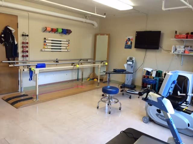 An interior rehabilitation room with parallel walking bars, exercise machines, stools, and a wall-mounted TV.