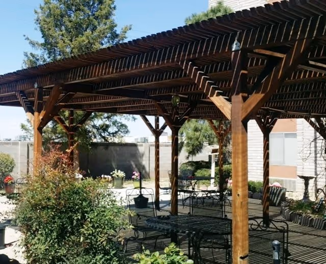 Outdoor patio area at Camino Retirement Apartments featuring a wooden pergola providing shade over metal tables and chairs, surrounded by plants and greenery with a building and trees in the background under a clear blue sky.