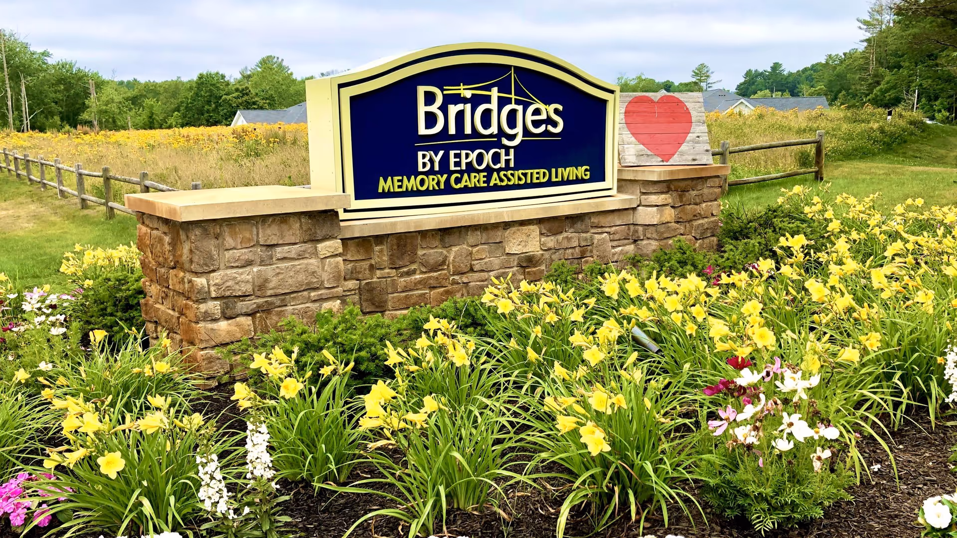 Stone sign for Bridges by EPOCH Memory Care Assisted Living surrounded by yellow and white flowers with a wooden fence and trees in the background under a cloudy sky.
