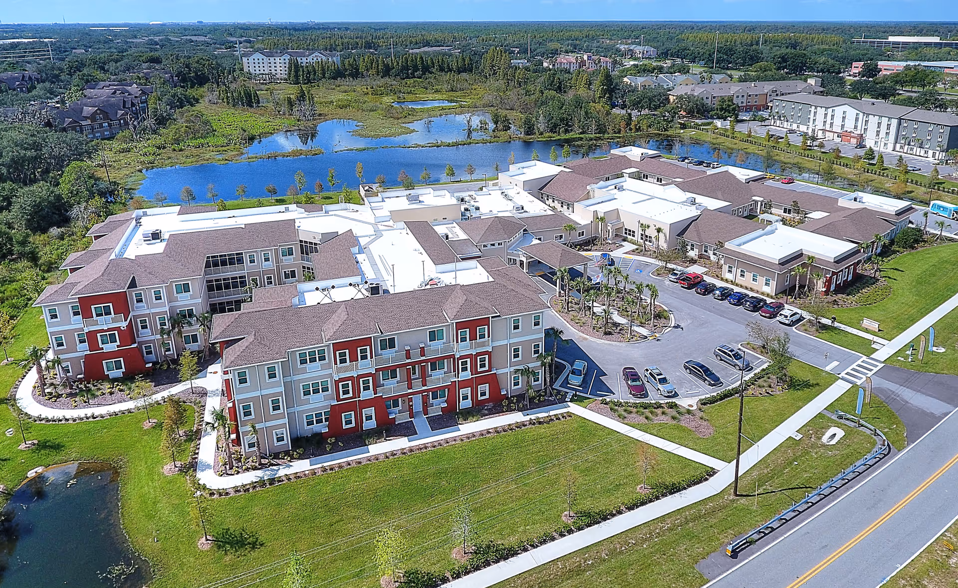 Aerial view of Promise Pointe Senior Living facility showing multiple connected buildings with red and beige exterior walls, surrounded by green lawns, parking lots with cars, and a nearby pond with trees and vegetation in the background under a clear blue sky.