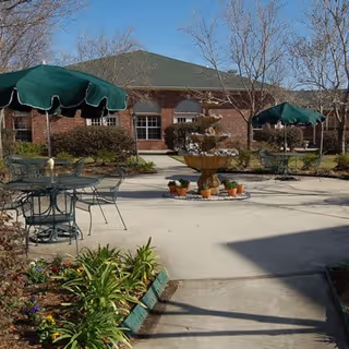 Courtyard with a central tiered fountain, metal tables with green umbrellas, flower beds, and a brick building in the background.