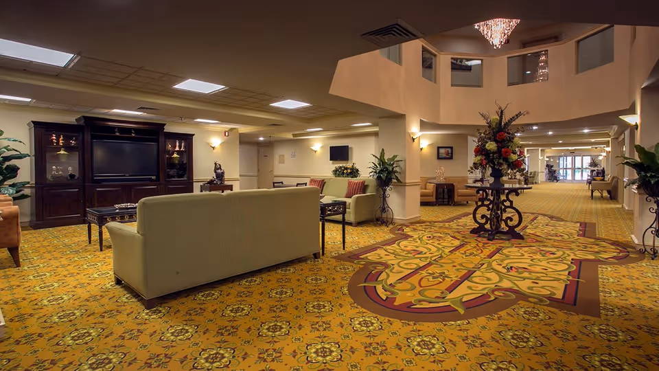 Spacious senior living community common area with patterned yellow carpet, beige sofas, dark wood entertainment center with a TV, decorative plants, and a round table with a large floral arrangement under a chandelier.