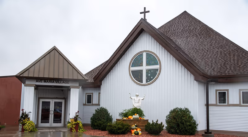 Front exterior of a white church-like building with a cross-topped gable, round stained-glass window, statue, landscaping and an entrance labeled "AVE MARIA VILLAGE".