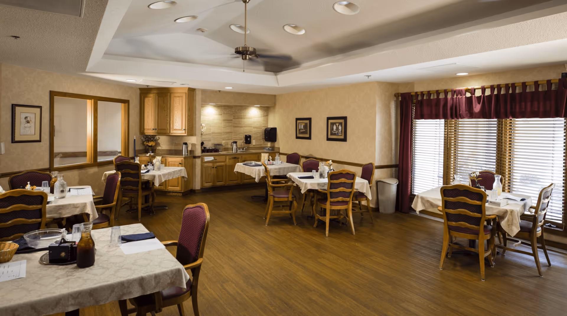 Dining room with several set tables and wooden chairs, a serving counter/kitchenette, and large windows with blinds and curtains.